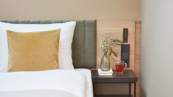 Una cama bien hecha con un cojín blanco y dorado, un cojín verde y una mesita de noche con un florero de flores y un vaso de bebida roja.