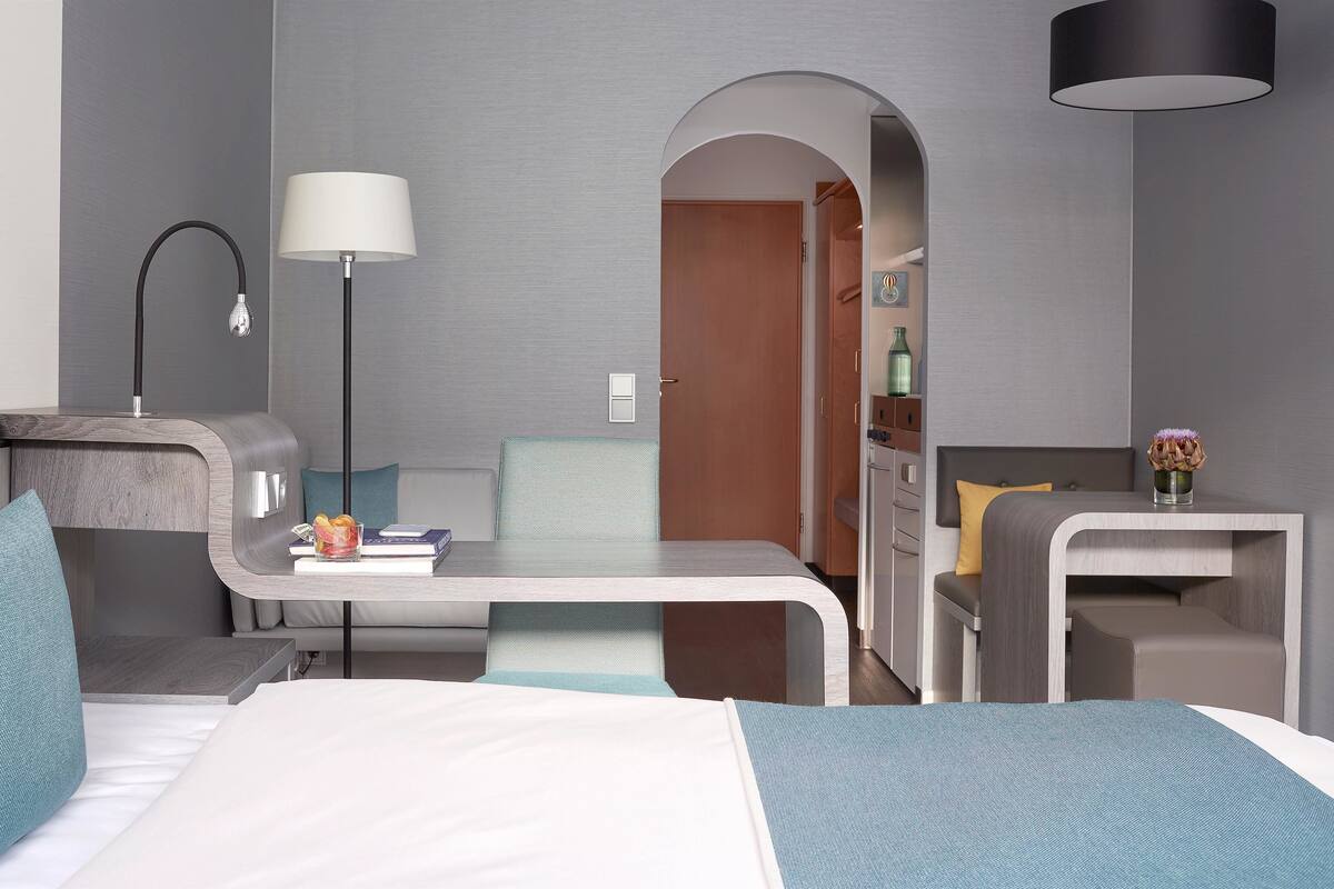 Ein modernes Hotelzimmer mit Bett, Schreibtisch, Sessel, Lampe und einem kleinen Tisch mit Blumen.