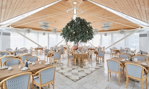 A spacious dining area with wooden tables and chairs, a large central tree, and a high ceiling with skylights. - Living Hotel Nürnberg (Nuremberg)