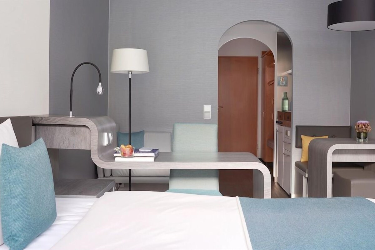 A modern hotel room with a bed, a desk, a chair, a lamp, and a small table with a vase of flowers.