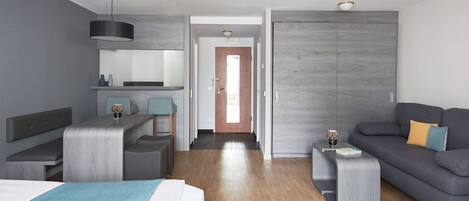 Ein modernes Hotelzimmer mit einem Bett, einer Couch, einem kleinen Tisch und einem Schreibtisch. Eine Tür führt zu einem Flur.