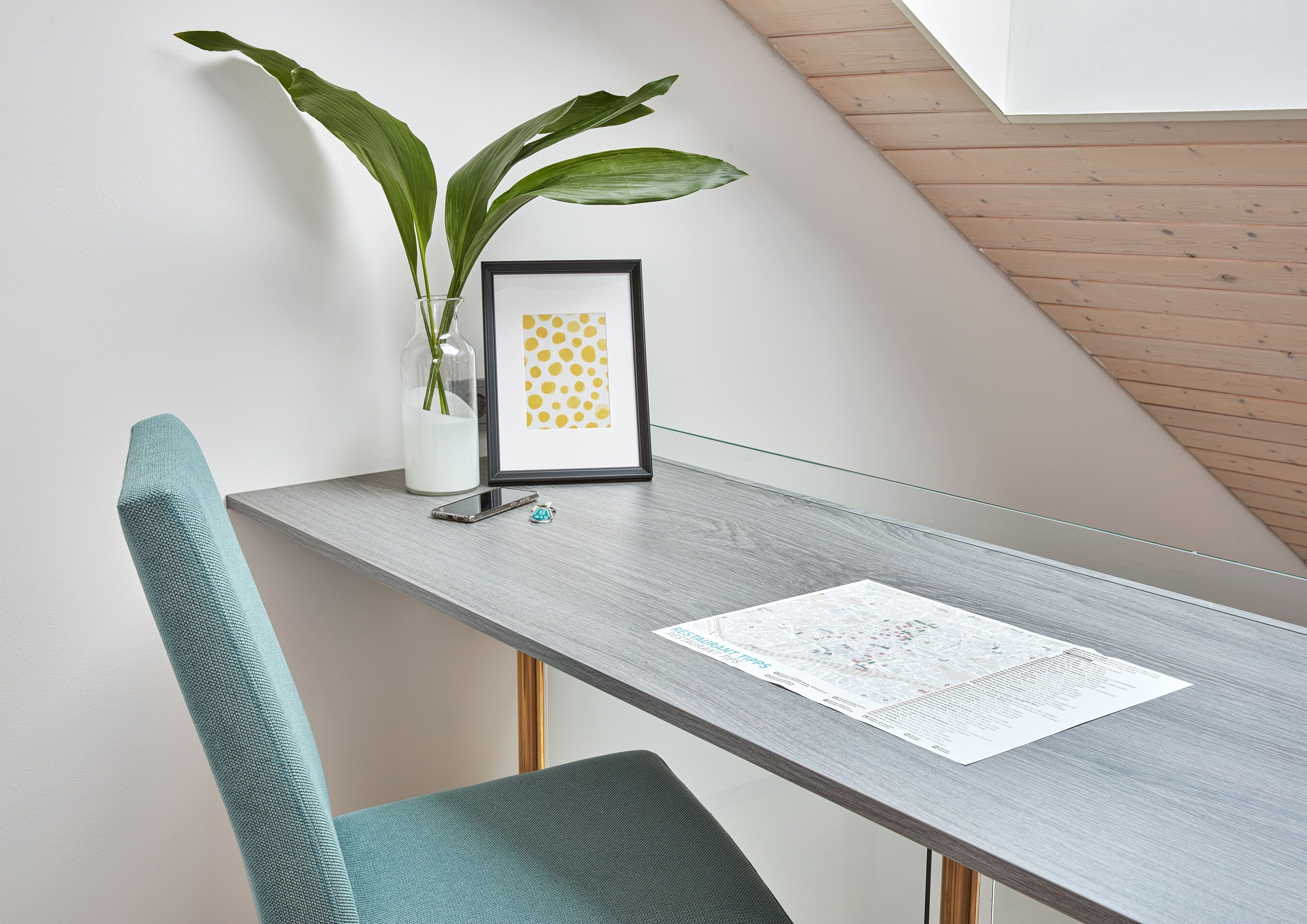 Una habitación moderna con techo inclinado de madera, una mesa gris con un cuadro enmarco, un florero con hojas verdes y una silla color turquesa.