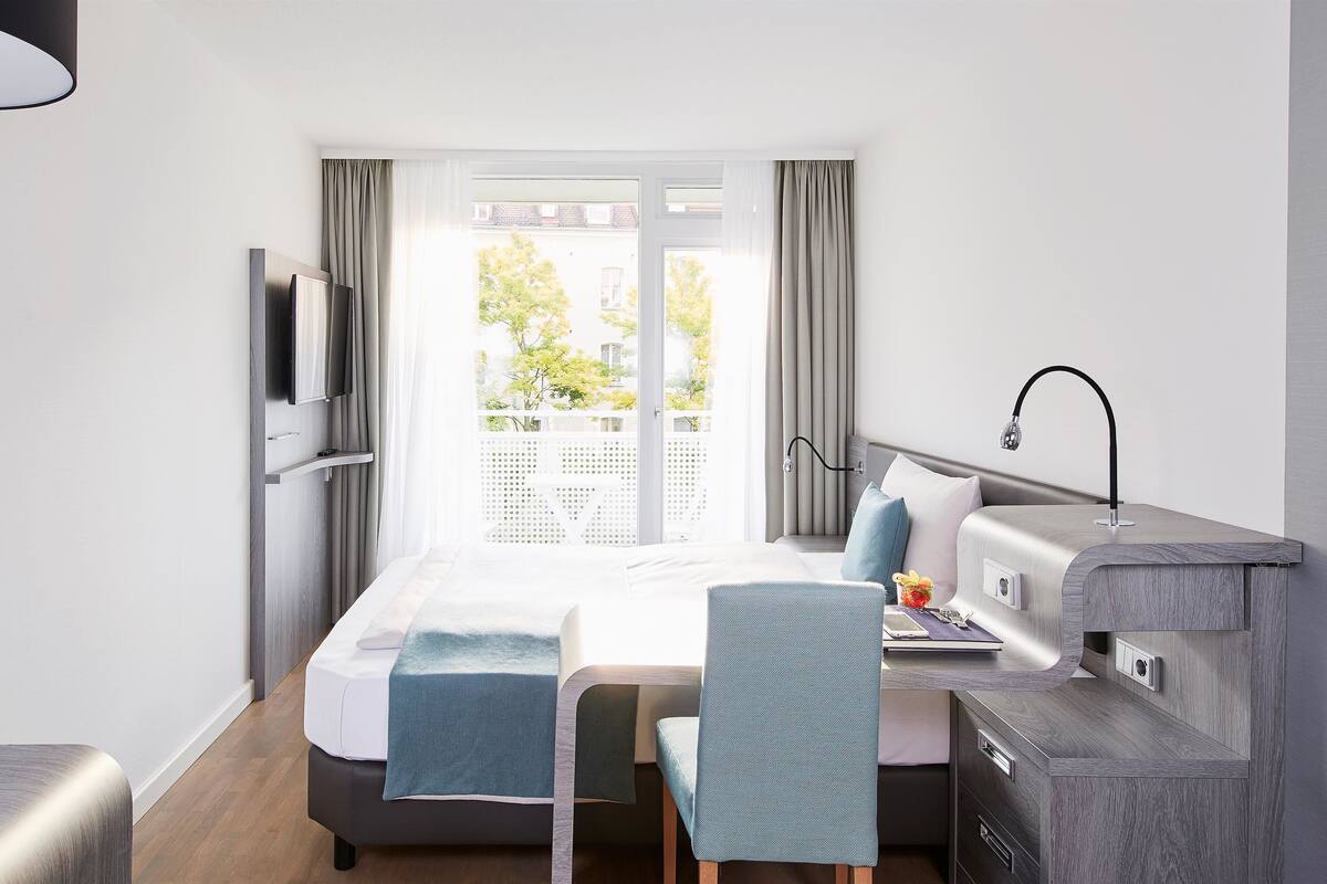 Ein modernes Hotelzimmer mit Bett, Schreibtisch, Stuhl, Fernseher und einem Balkon mit Vorhängen.