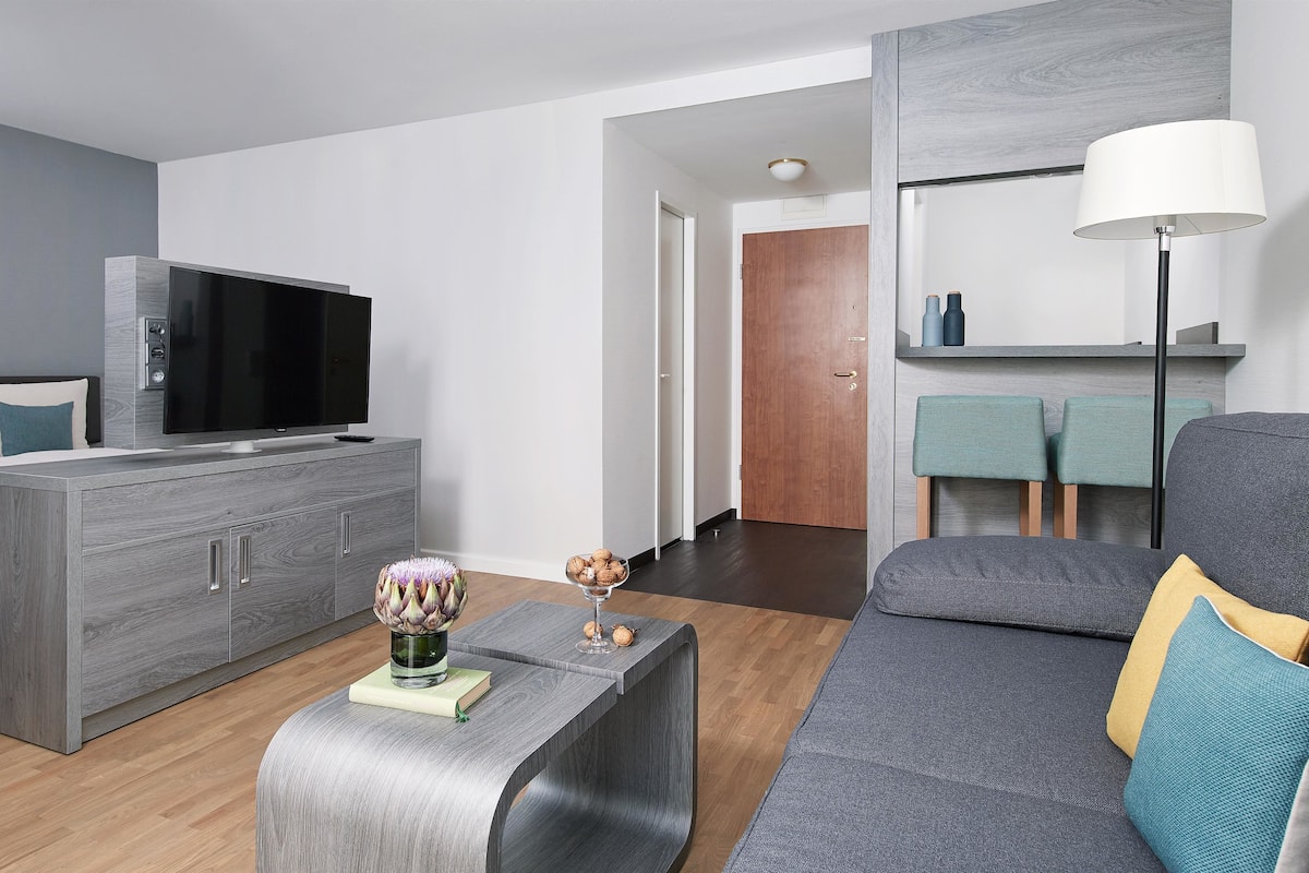 Ein modernes Wohnzimmer mit einem grauen Sofa, einem Fernseher auf einem Holzschrank, einem kleinen Tisch mit einer Vase und einer Lampe.