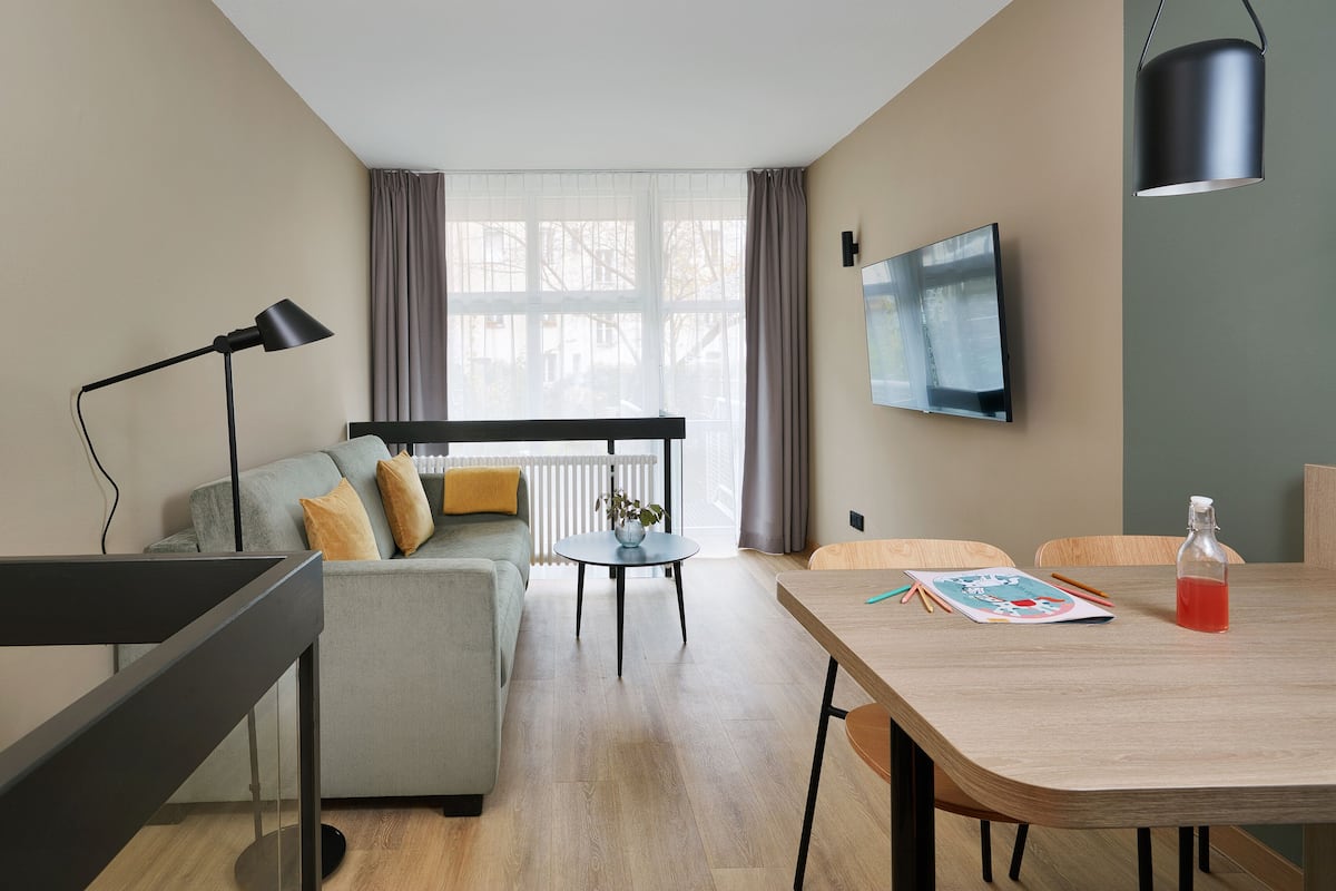 Ein modernes Wohnzimmer mit einem grauen Sofa, einem runden Tisch mit einer Vase, einer schwarzen Stehlampe und einem Holzessttisch mit einer Flasche und Unterlagen.