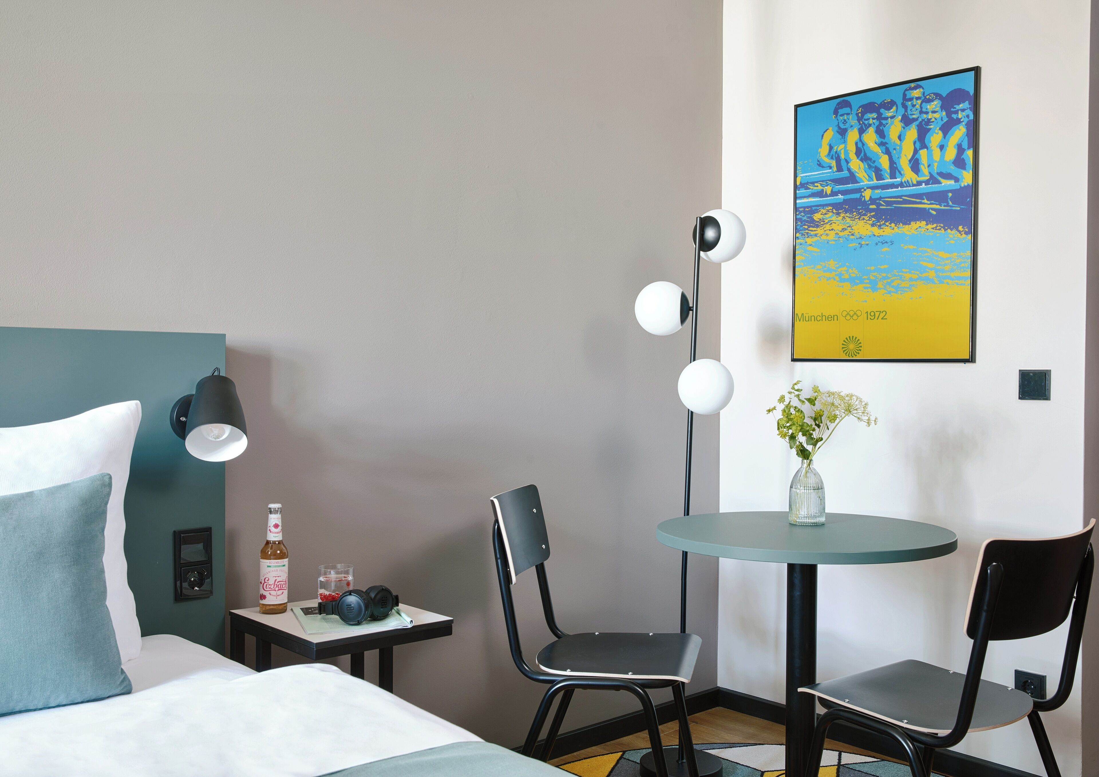Une chambre d’hôtel avec un lit, une petite table sur laquelle se trouvent une bouteille et un casque audio, deux chaises, une table ronde avec un vase, et une affiche accrochée au mur.