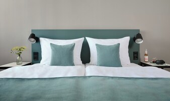 Cama bem arrumada com roupa de cama branca e azul-turquesa, dois travesseiros combinando, um vaso com flores amarelas, uma garrafa de cerveja e um par de fones de ouvido em uma mesa de cabeceira.