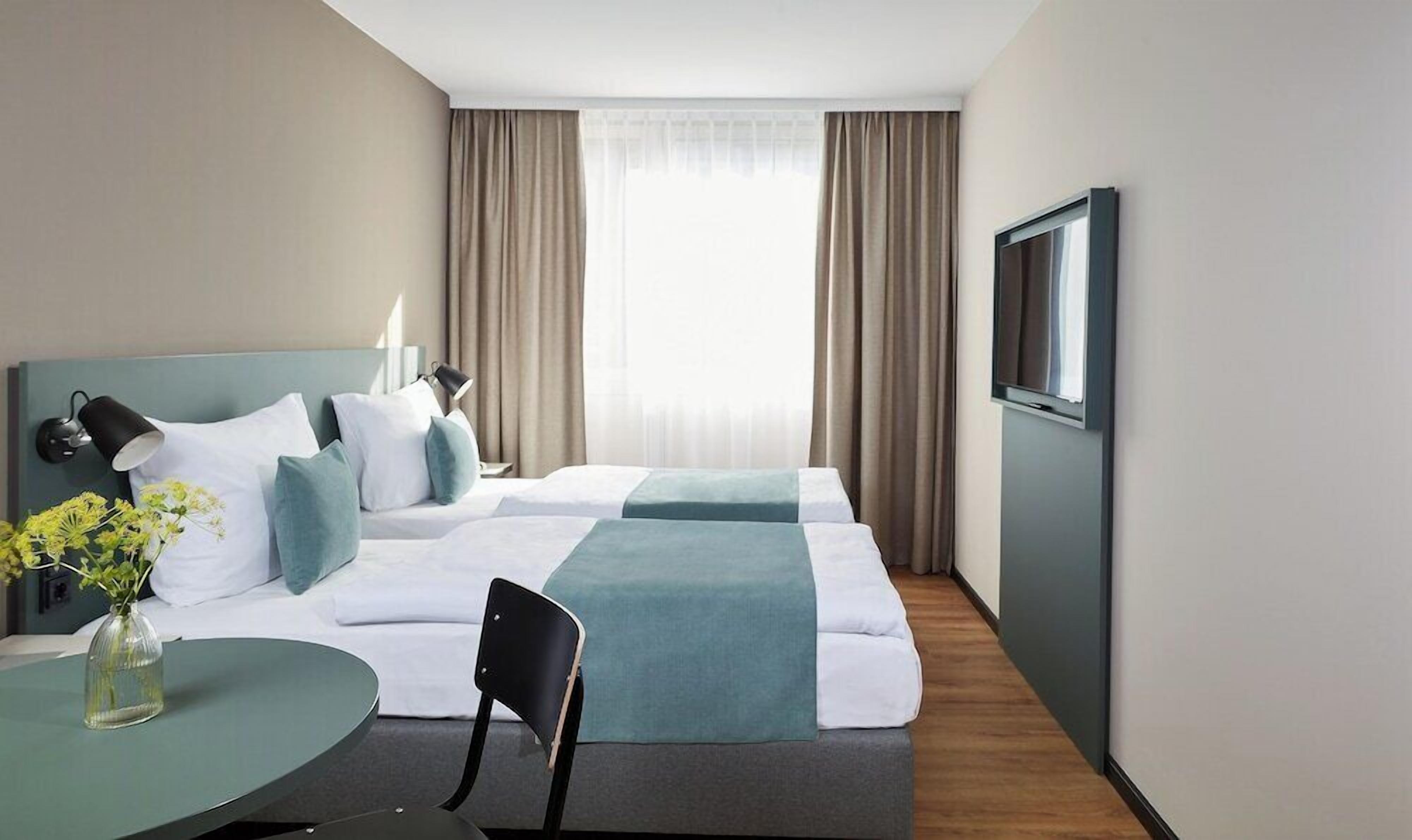 A hotel room with a bed, two bedside tables, a chair, a TV, and a window with curtains.