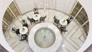 Uma área de refeições circular com piso de mármore e cadeiras pretas ao redor de mesas brancas, com uma fonte central.