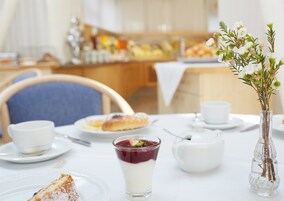 Uma mesa de jantar com um prato de sobremesa, um copo de sobremesa, uma xícara, um vaso com flores e uma cadeira.