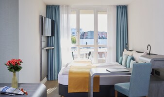 Quarto de hotel com uma cama, escrivaninha, cadeira, televisão e janela com cortinas.