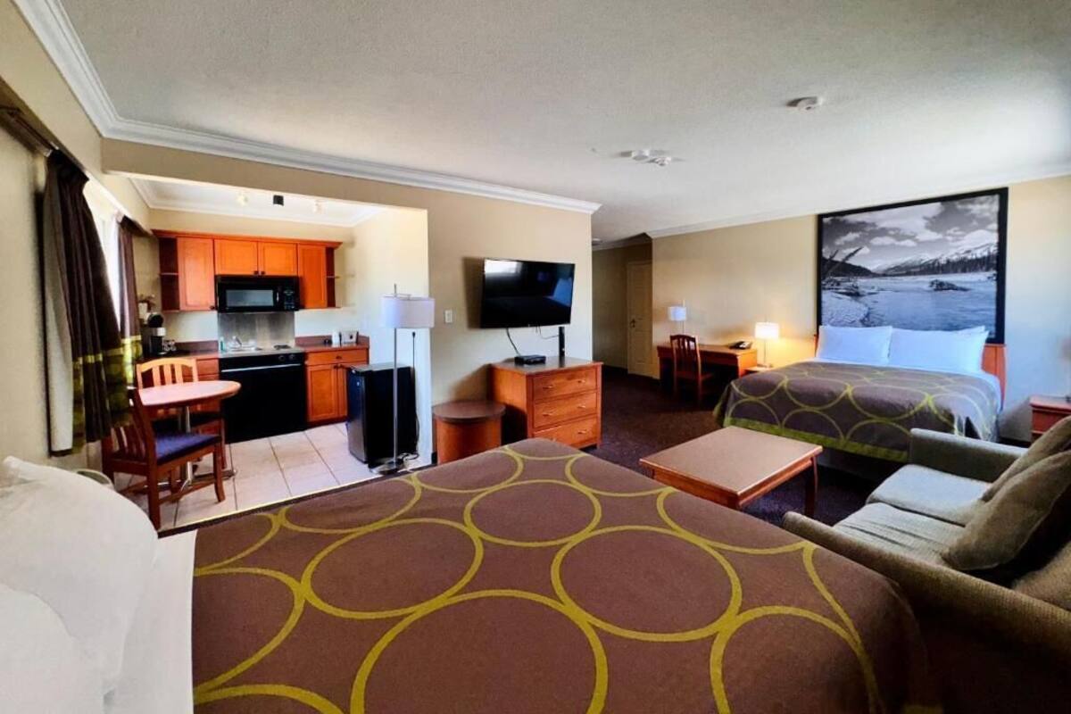 A hotel room with two beds, a kitchenette, a TV, and a painting of a mountain landscape.