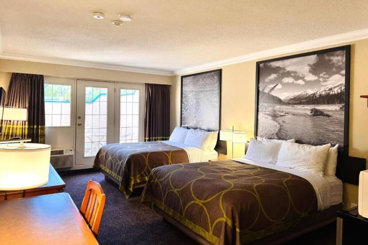 A hotel room with two beds, a desk, a chair, a window with curtains, and a large framed picture on the wall.