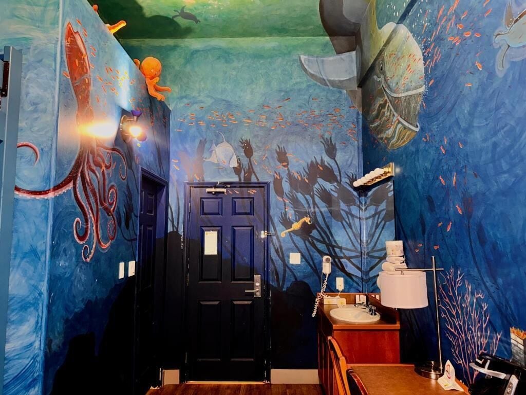 Una stanza con un affresco subacqueo, una porta nera e un comò in legno con uno specchio.