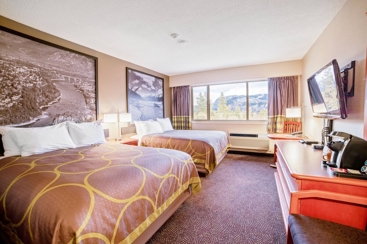 A hotel room with two beds, a desk with a coffee maker, a TV, and large windows with curtains.