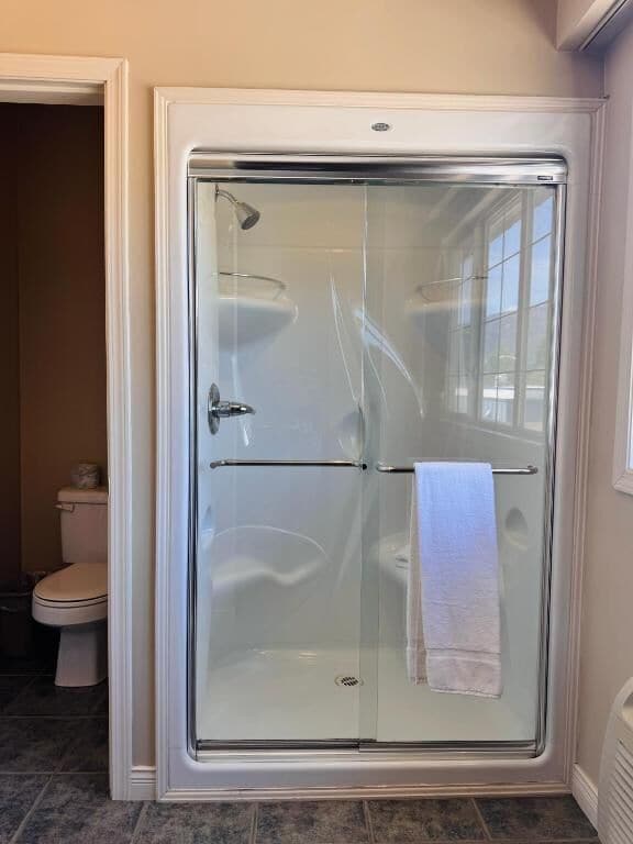 Une douche fermée par des parois en verre, avec une serviette accrochée à la porte, des toilettes visibles sur la gauche et un sol carrelé.
