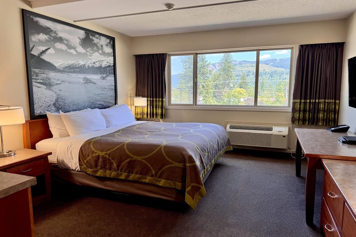 A hotel room with a bed, a desk, a chair, a window with a mountain view, and a large landscape painting on the wall.