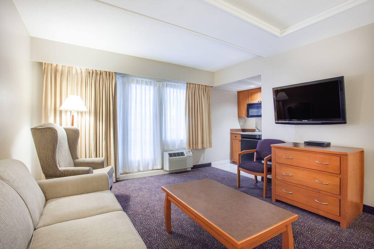 A hotel room with a sofa, armchair, wooden table, TV, and kitchenette.