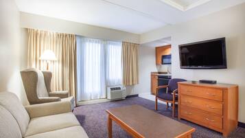 A hotel room with a sofa, armchair, wooden table, TV, and kitchenette.