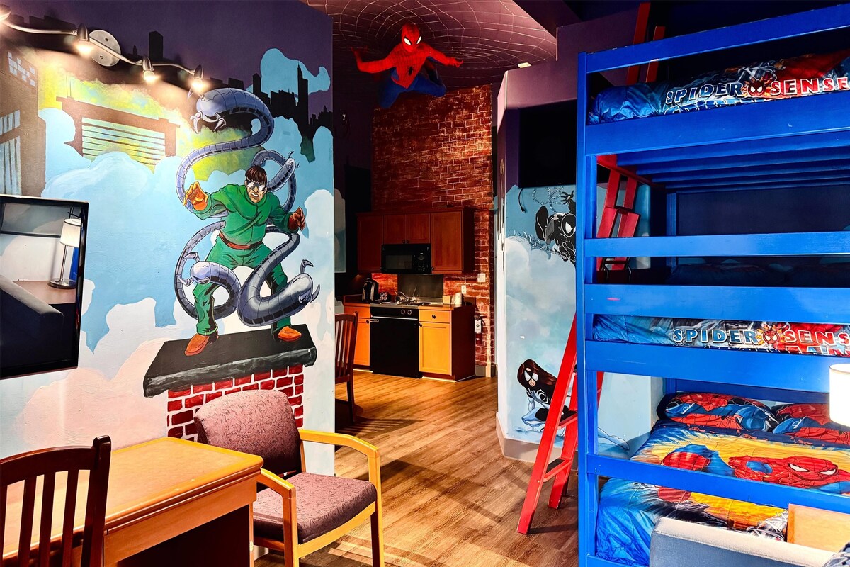 A room with a mural of Spider-Man, a bunk bed with Spider-Man bedding, a wooden table, and a kitchen area in the background.