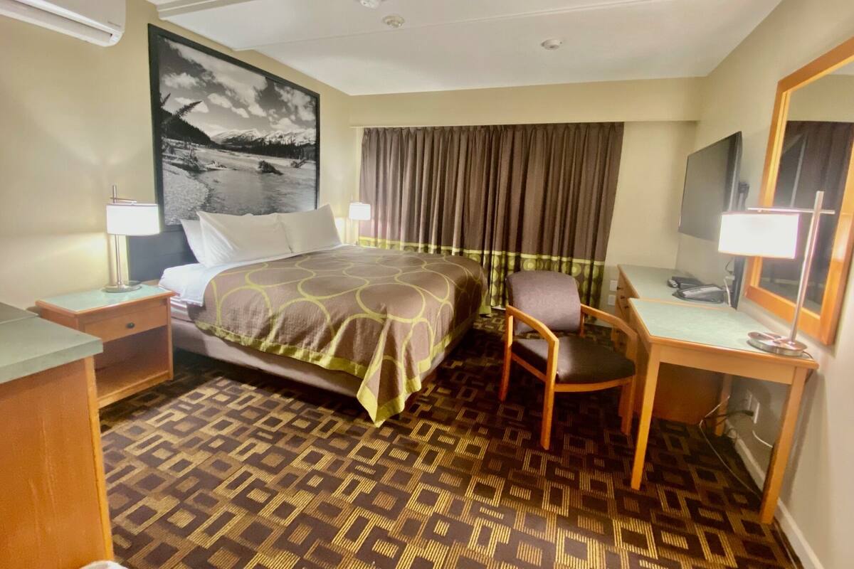 A hotel room with a bed, a desk, a chair, a desk lamp, a television, and a patterned carpet.