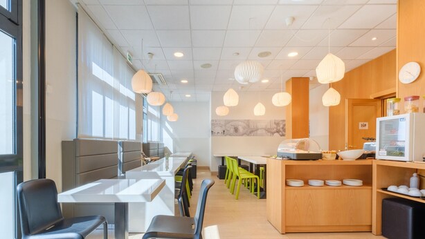 A modern dining area with a long communal table, individual seating, and a kitchen counter with a refrigerator and food display. - B&B Hotel Ravenna (Ravenna)