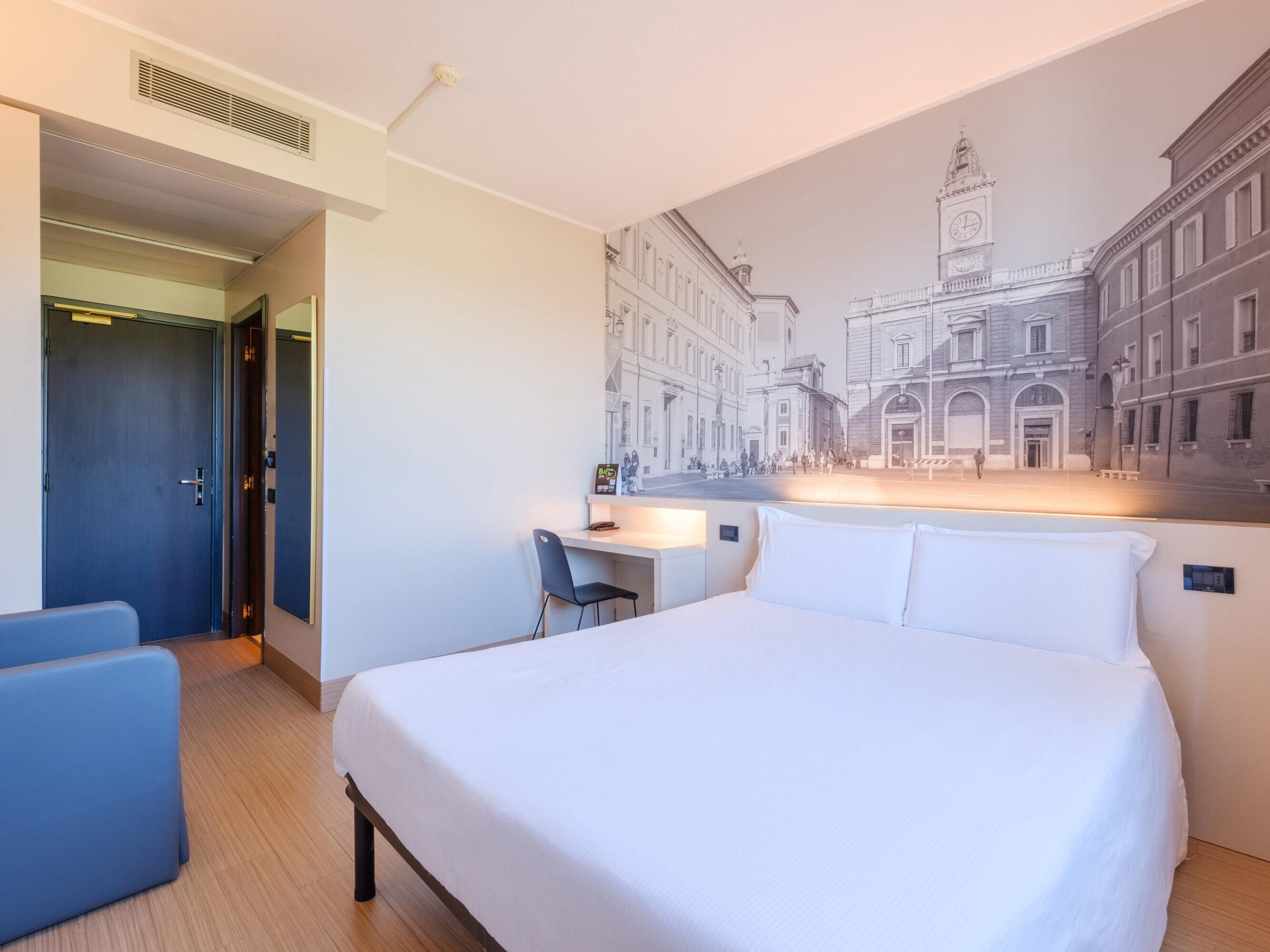 Camera d'albergo con un letto, una scrivania e una sedia. Una grande parete decorata con un affresco raffigurante un paesaggio urbano.