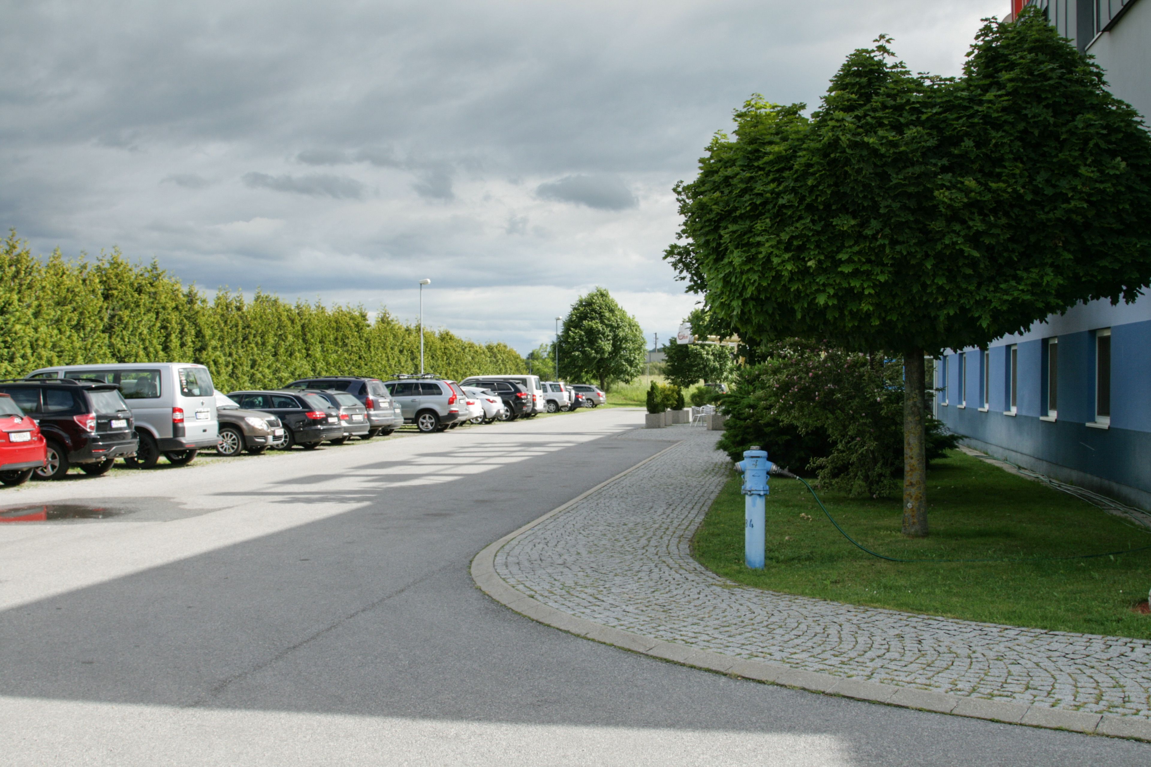 Un parking où de nombreuses voitures sont garées en rangées, un chemin pavé et un bâtiment à la façade bleue, situé sur la droite.
