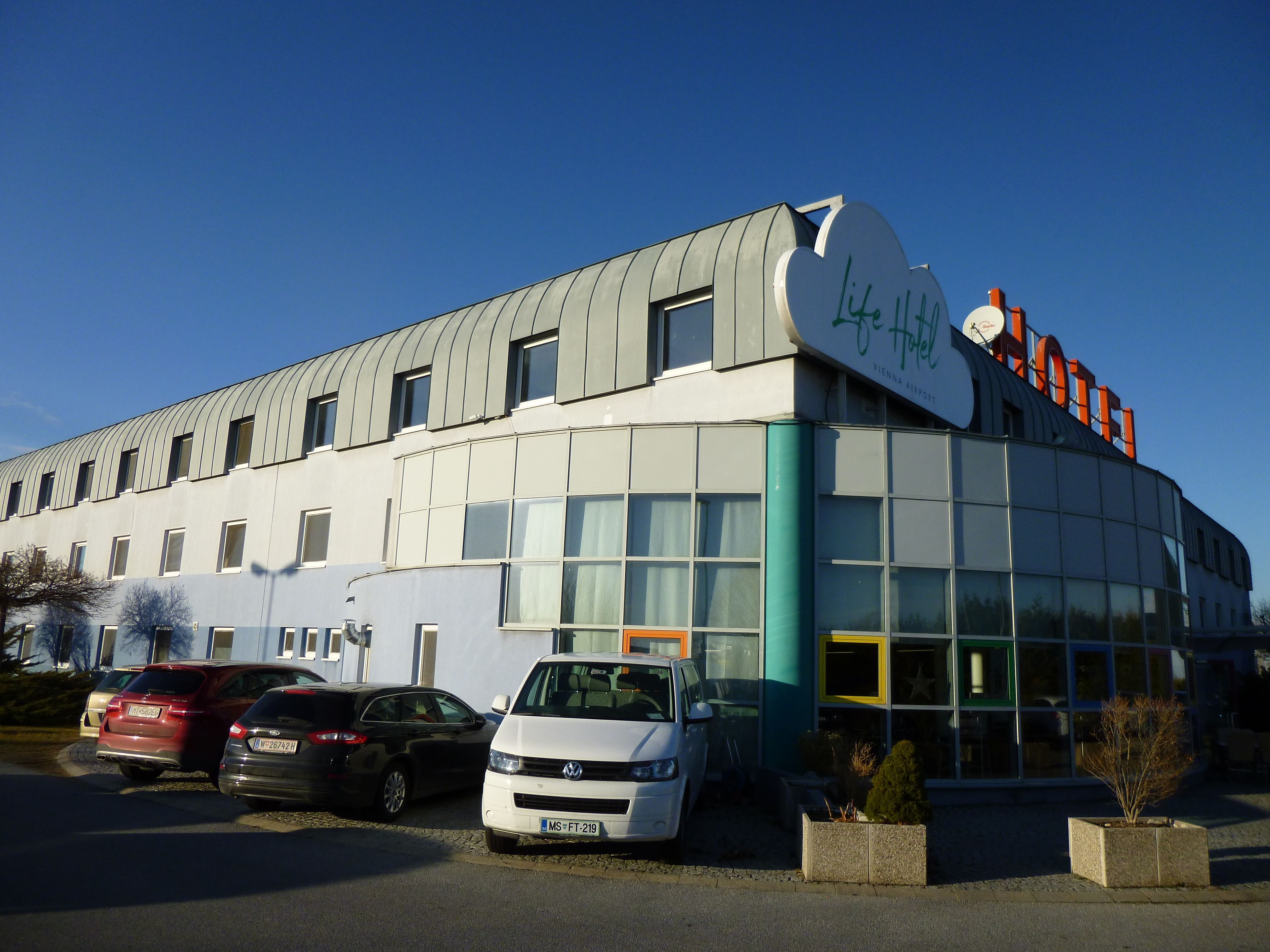 A modern hotel with a curved roof, large windows, and a sign indicating it is near Vienna Airport.