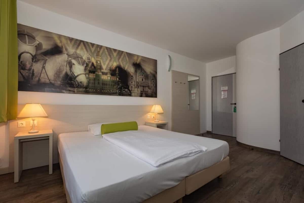 A hotel room with a large bed, two bedside tables with lamps, a green headboard, and a large painting on the wall.