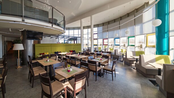 A spacious indoor dining area with multiple tables and chairs, a green accent wall, and large windows allowing natural light. - Life Hotel (Fischamend)