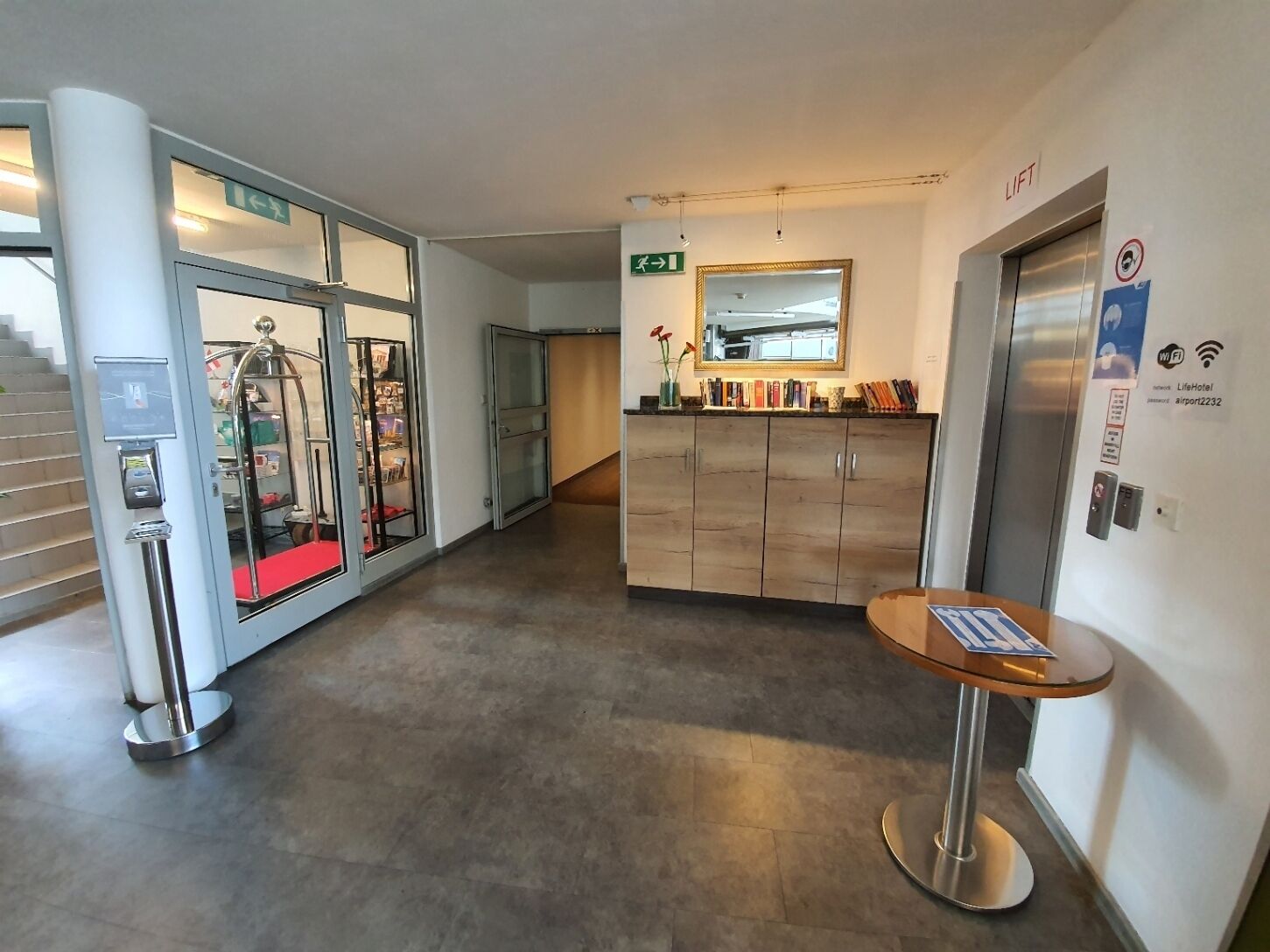 Un moderno vestíbulo de hotel con mostrador de recepción, puerta de cristal, ascensor y una pequeña mesa con un cartel.