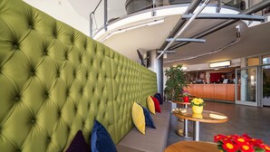 A green tufted sofa with colorful cushions, a wooden table with a yellow flower vase, and a reception area in the background. - Life Hotel (Fischamend)