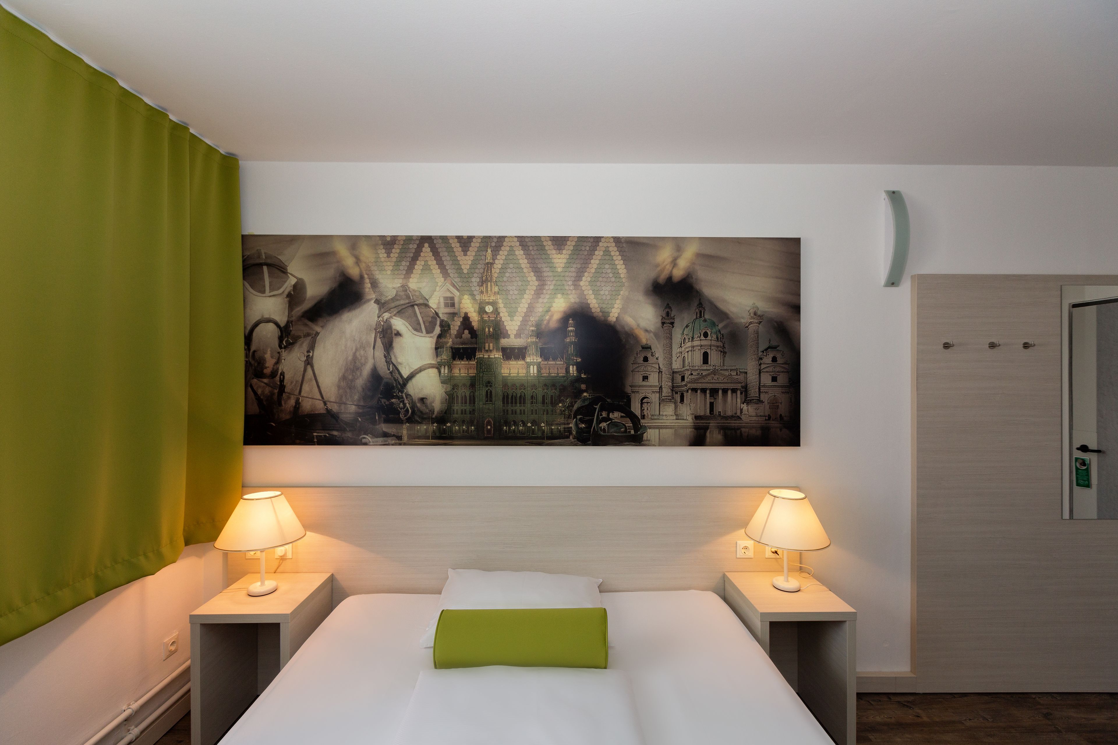 Habitación de hotel con una cama, dos lámparas de noche, un almohadón verde y un cuadro grande con una vista panorámica de la ciudad.