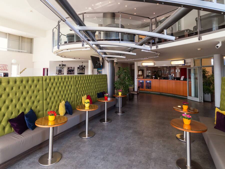 Un hall d'entrée moderne avec un espace salon agrémenté de sièges rembourrés verts, des tables rondes ornées de vases de fleurs et une rambarde en verre donnant sur l'ensemble de l'espace.