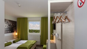 A modern hotel room with a bed, green curtains, a view of the outdoors, a small table with a lamp, and a coat rack. - Life Hotel (Fischamend)