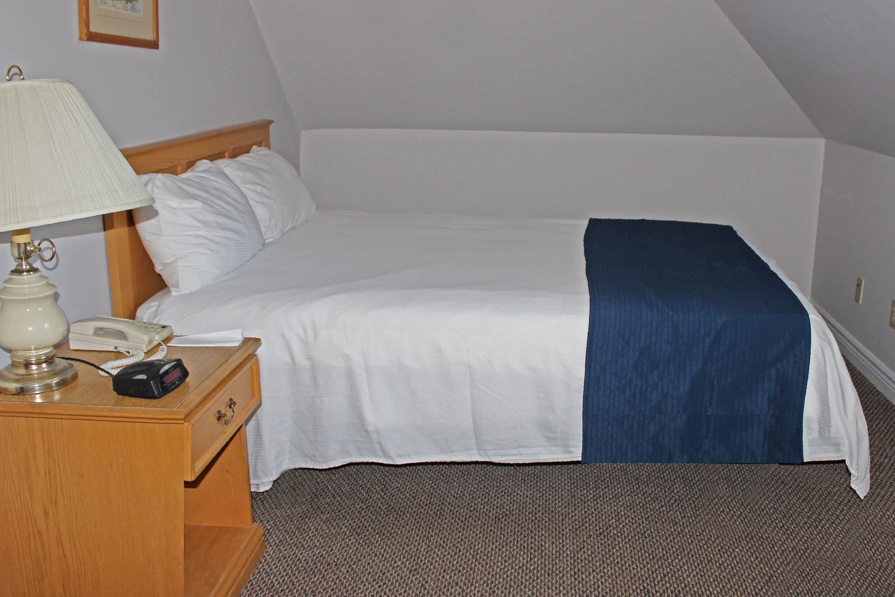 A hotel room with a bed, a nightstand with a lamp, a phone, and a digital clock.