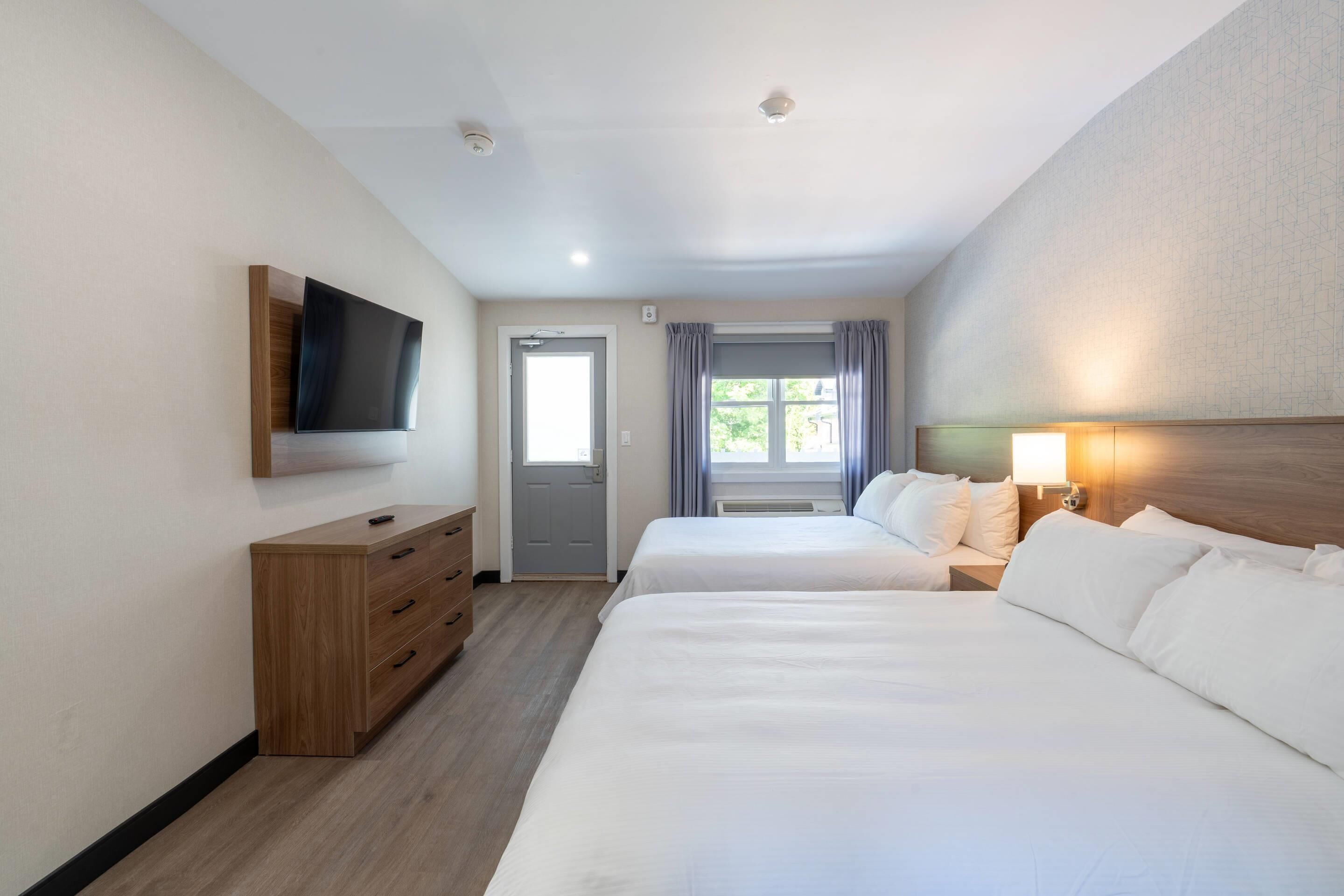 Habitación de hotel con dos camas, un televisor montado en la pared, un aparador de madera, una ventana con cortinas y una mesita de noche con lámpara.
