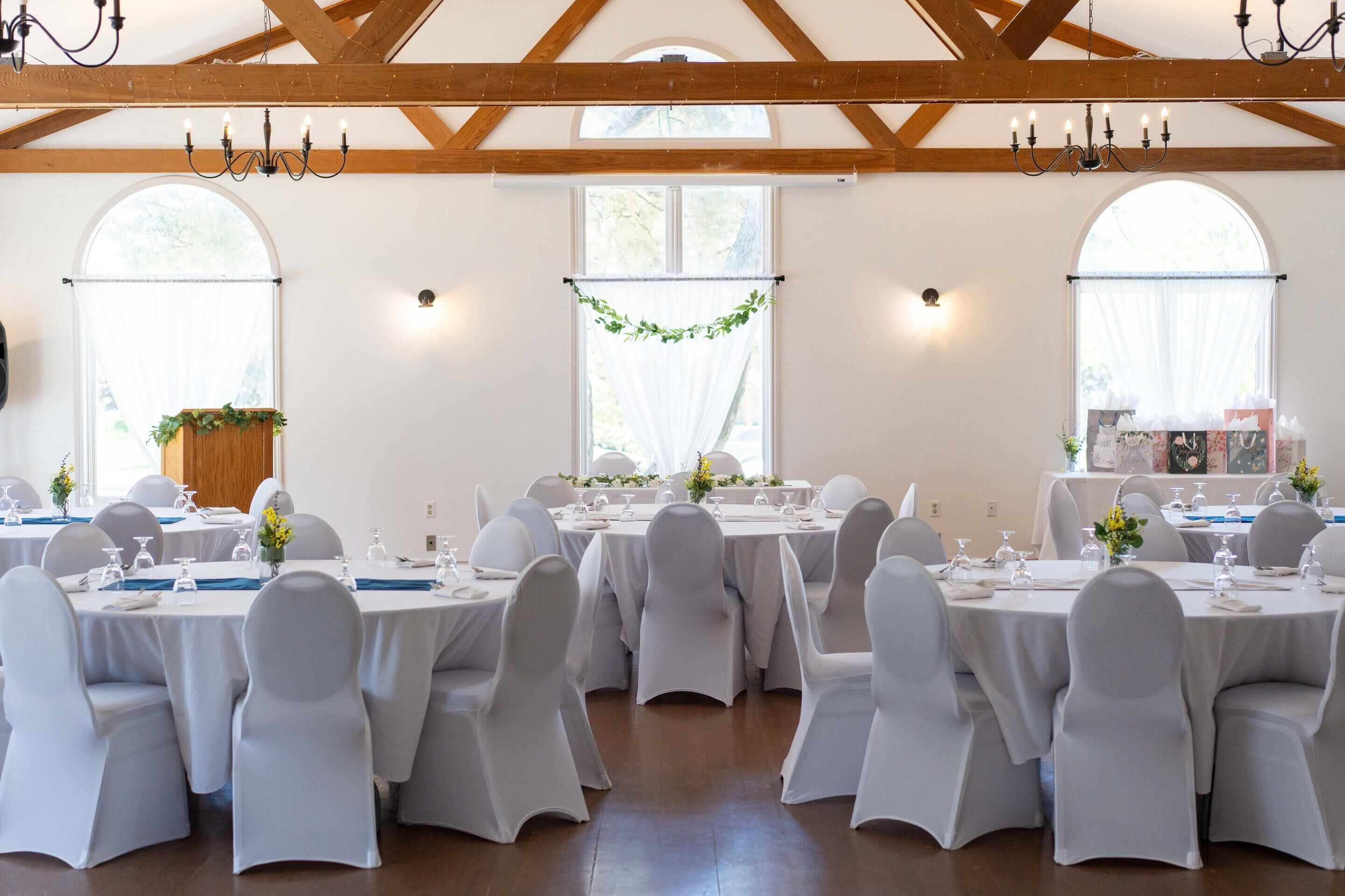 A banquet hall with round tables, white chairs, chandeliers, and large windows.