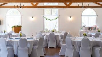 A banquet hall with round tables, white chairs, chandeliers, and large windows.