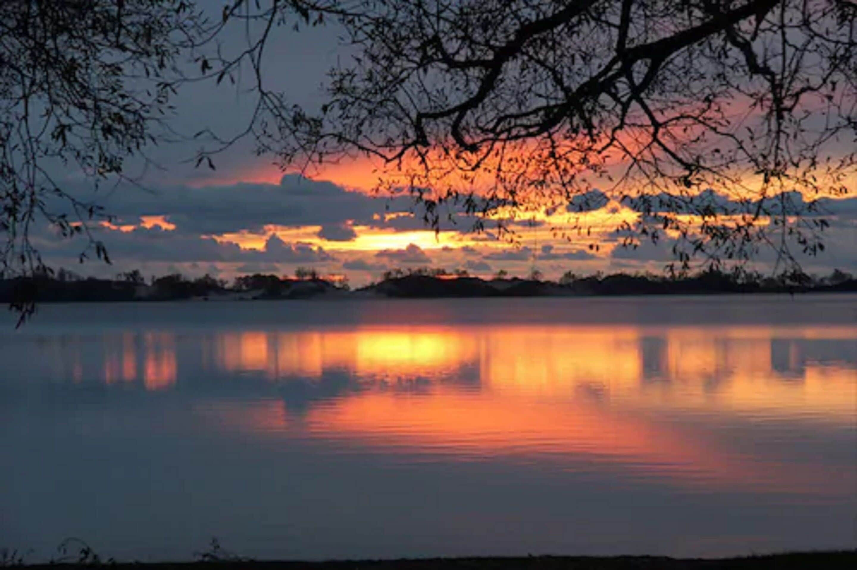 A calm lake reflecting a sunset with silhouettes of trees and distant hills.