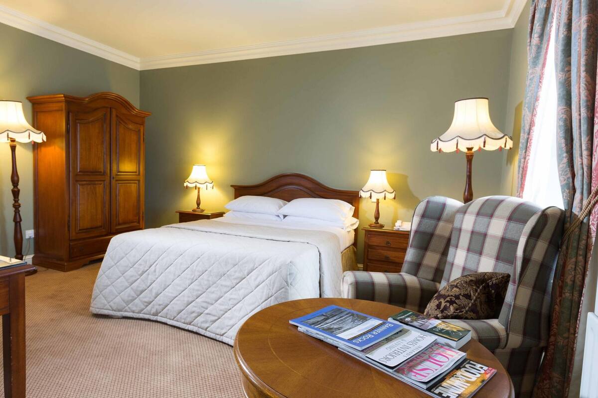 A hotel room with a bed, two bedside lamps, a wooden wardrobe, a plaid armchair, a small table with magazines, and a carpeted floor.