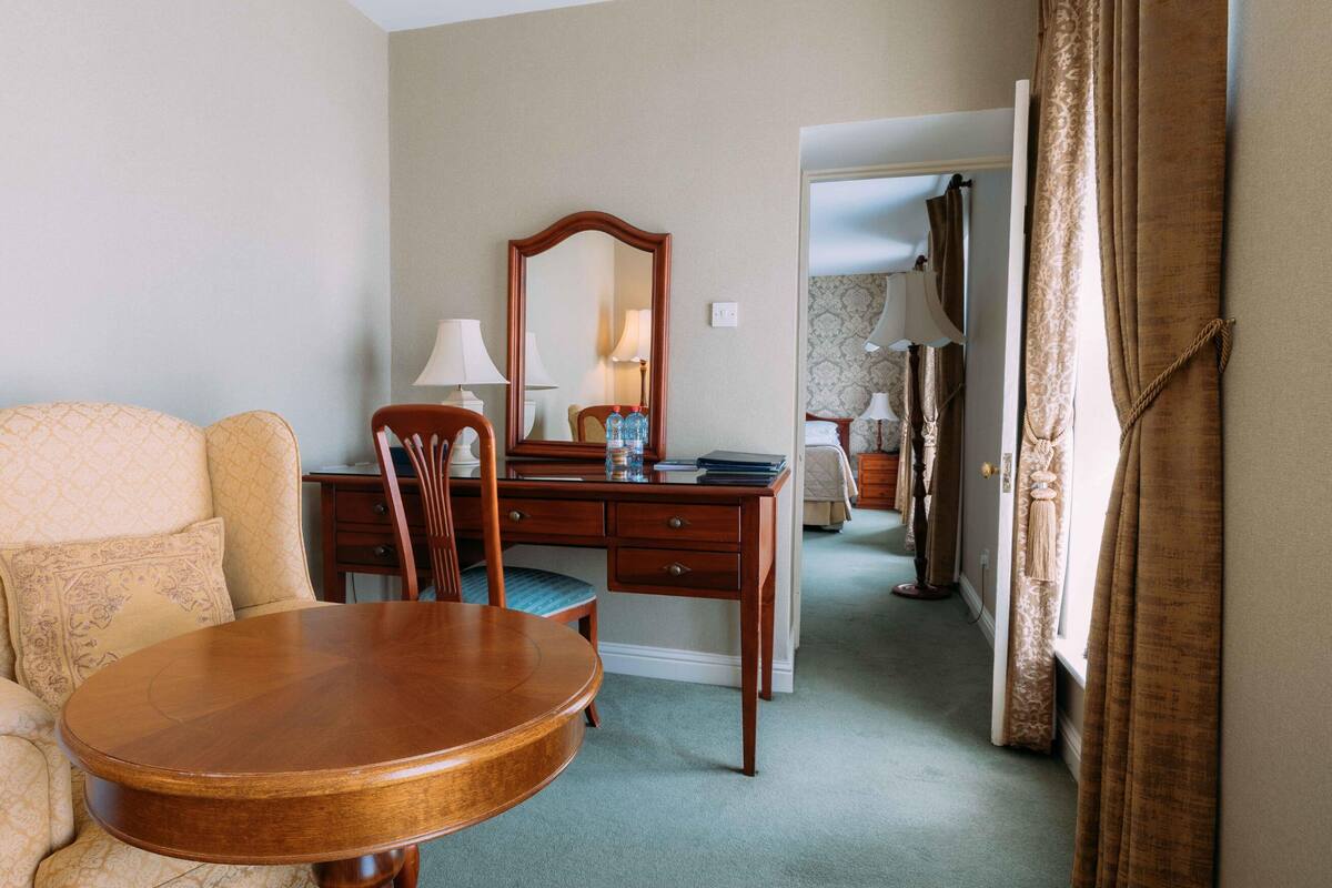 A hotel room with a wooden desk, a chair, a round table, a mirror, and a bed with a nightstand.