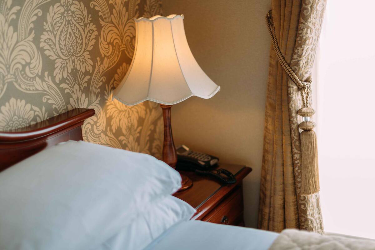 A neatly made bed with a white pillow and a wooden headboard, a bedside table with a lamp, and a patterned wallpaper.
