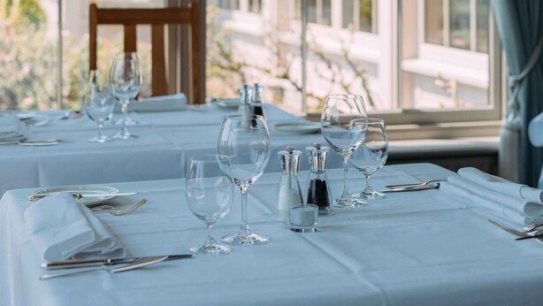 A dining table set with white tablecloths, glasses, and cutlery, with a view of a building through large windows. - Sheedy's Country House Hotel (Lisdoonvarna)