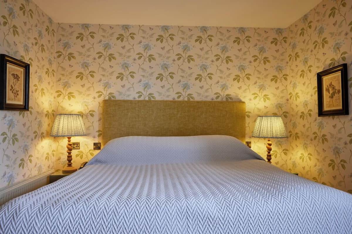A neatly made bed with a patterned bedspread, flanked by two bedside lamps on a nightstand, set against a floral wallpaper backdrop.