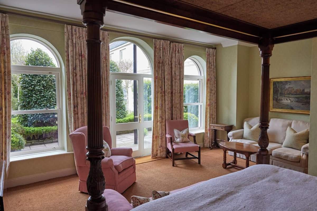 A room with a four-poster bed, two armchairs, a sofa, and a table. Large windows with curtains provide views of greenery outside.