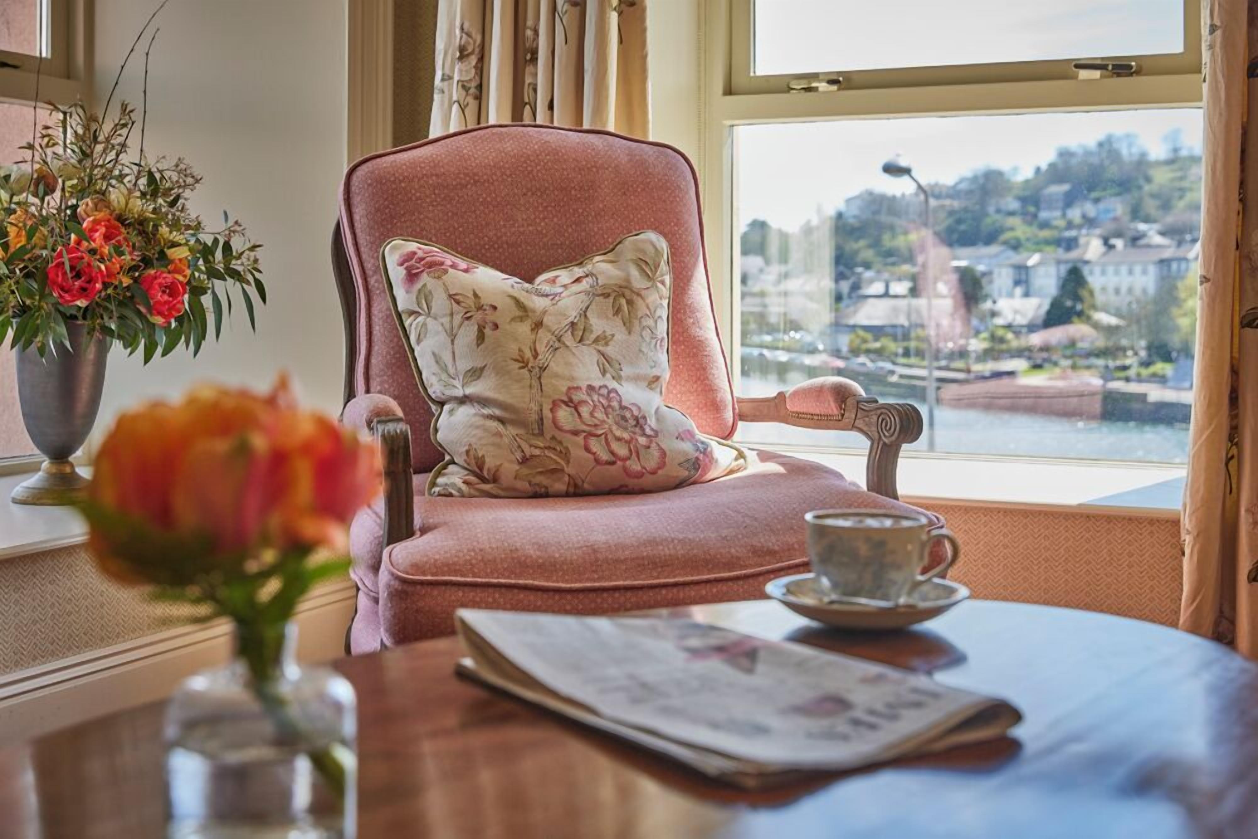 A room with a pink armchair, a floral pillow, a vase with flowers, a newspaper, and a cup on a table.