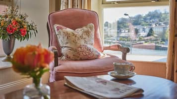 A room with a pink armchair, a floral pillow, a vase with flowers, a newspaper, and a cup on a table.