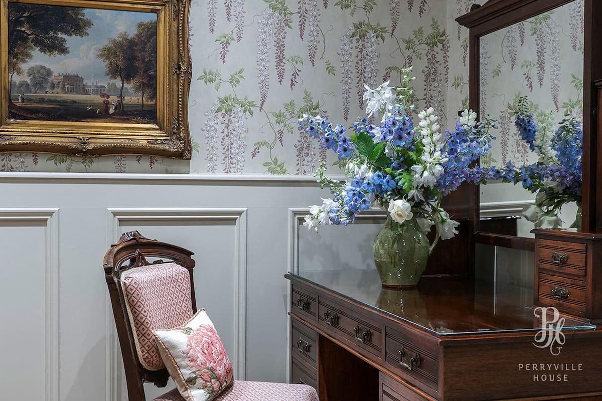 A room with a wooden dresser, a chair with a floral cushion, a vase of flowers, and a framed painting on the wall.